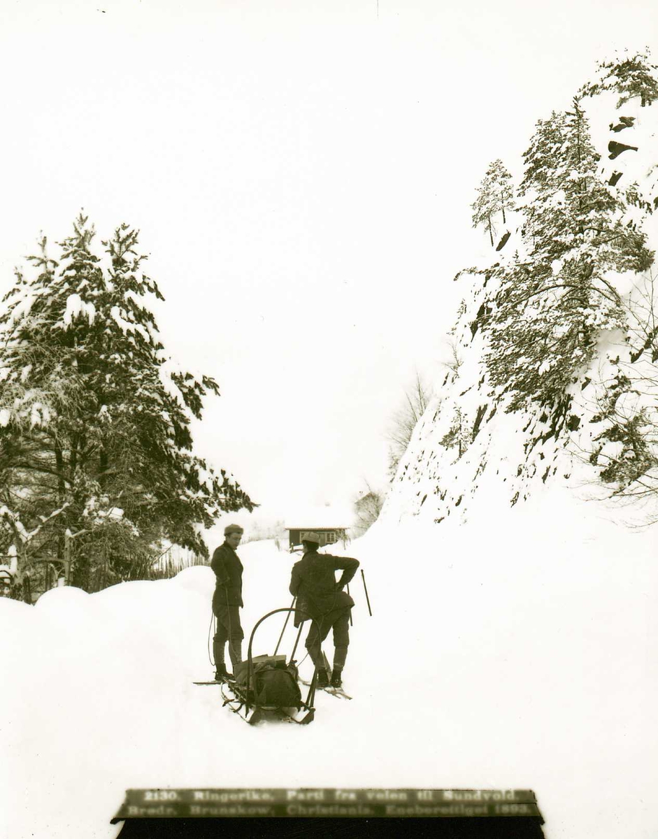Parti fra veien til Sundvollen, Hole, med to menn på ski hvilende ved siden av slede med oppakning. Snørikt landskap med et hus i bakgrunnen. Ant. fotografene Brødrene Brunskow. Del av deres serie norske lanskapsbilder.