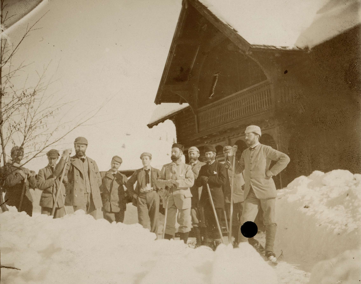 En gruppe menn måker snø foran trebygning, ukjent sted.