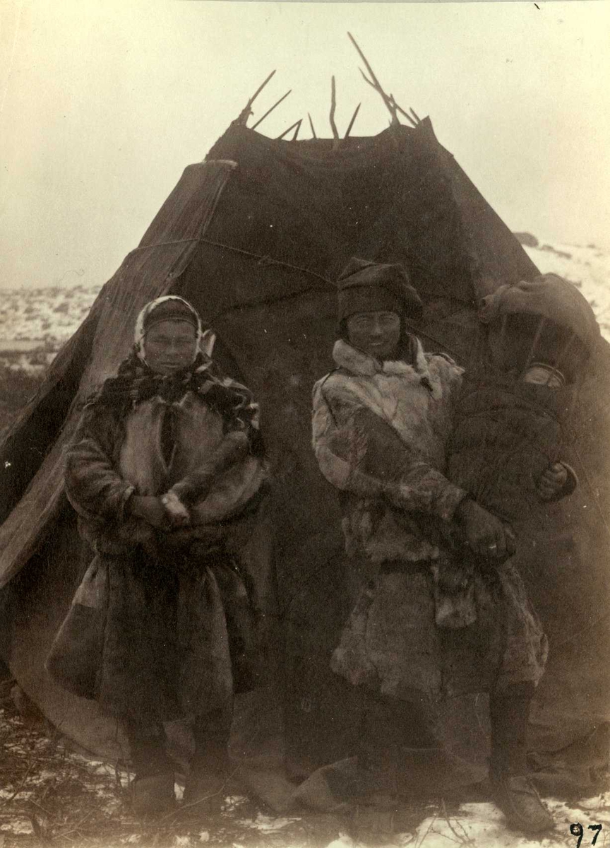 Norge. Vinterbilde av mann og kvinne i samisk drakt med barn i komse, står foran telt, lavo.