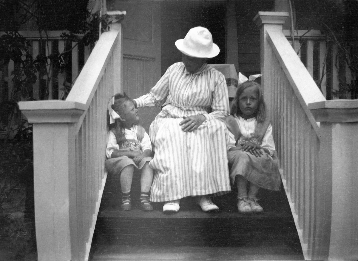 En kvinne sitter mellom to barn på en trapp. Kvinnen klapper den ene jenta på hodet. Robsahm og Lund.