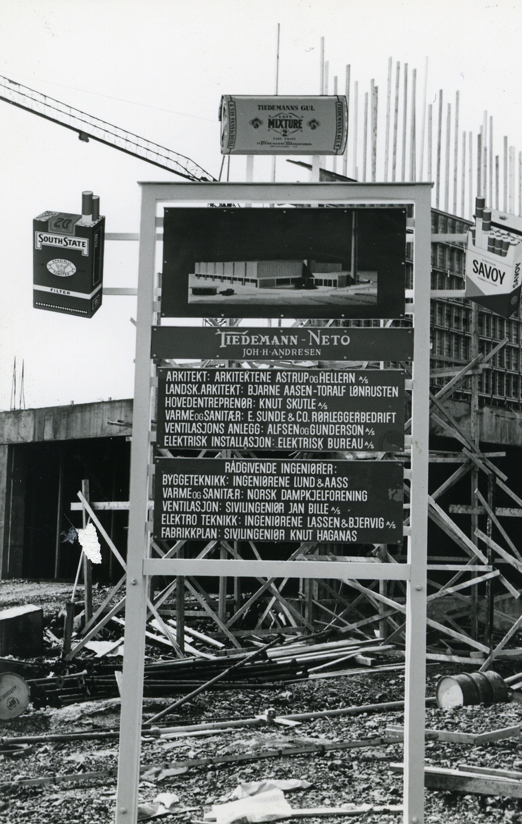 Byggeplass. 
Konstruksjon av Tiedemanns Tobaksfabrik på Hovin i 1967. Fotografiet viser en skilt på byggeplassen med informasjon om byggeprosjektet og reklamebilder for Tiedemanns tobakk og sigaretter.
