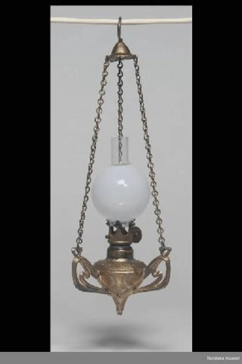 Inventering Sesam 1996-1999:
H 9,5 cm
B 5,5 cm
Taklampa, föreställande fotogenlampa. Oljehus med tre genombrutna armar av metall målat med guldfärg. Brännare med reglage och veke av tråd. Lampan hänger i kedjor, fästa i de tre armarna och förenade upptill i en klockliknande hållare med hängögla. Rund (lös) lampkupa av vitt glas med brännglas.
Har tillhört givarens mor och moster, Astrid (f 1903) och Kerstin (född 1908), döttrar till fabrikör Axel Reuterberg, Göteborg. Denne köpte leksaker på affärsresor, bl a i Tyskland.
I A-Berg nov 73/= Inga Arnö-Berg/
Leif Wallin maj 1997