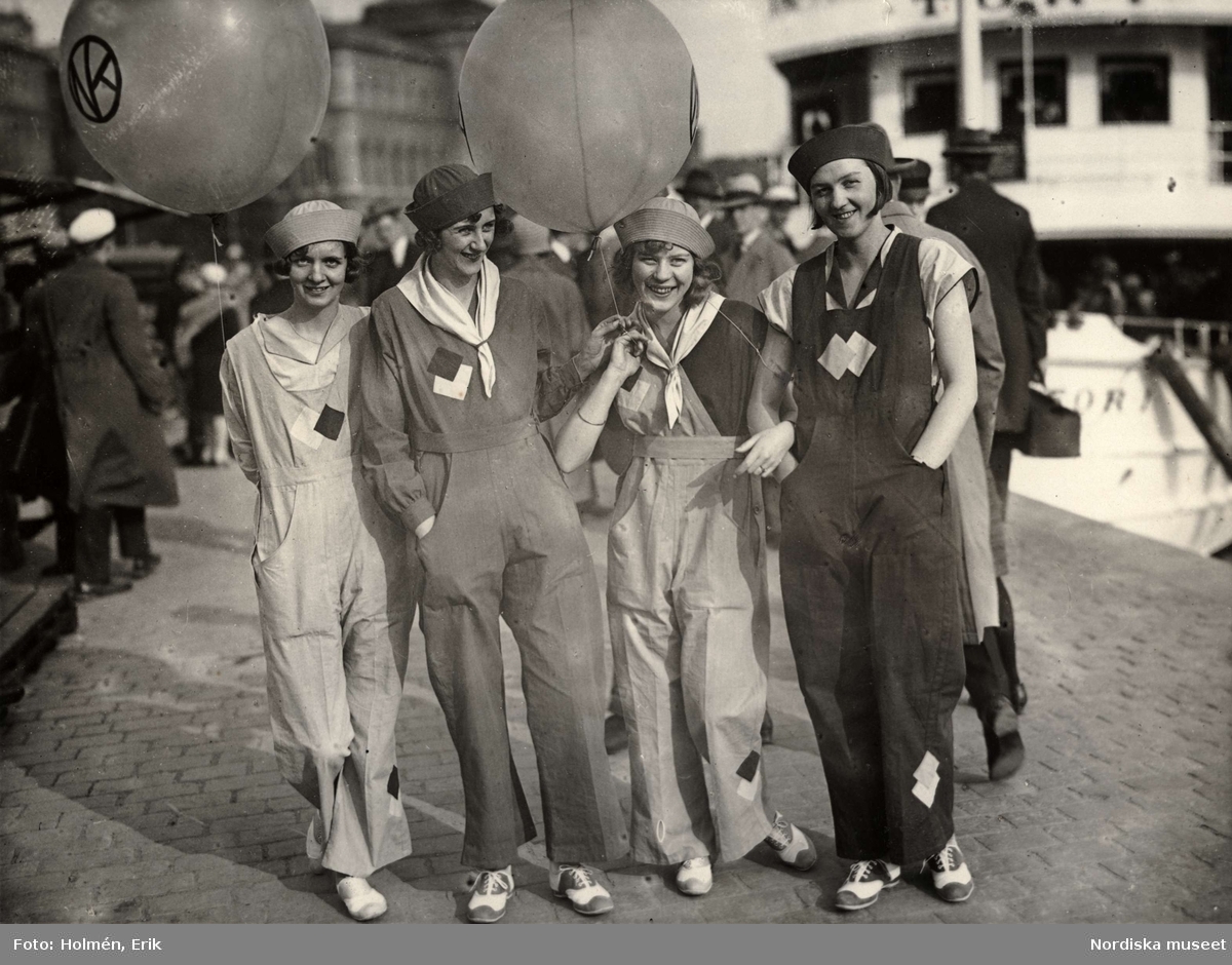 Fyra kvinnliga modeller iklädda overaller, håller i två reklamballonger. "NK (Nordiska Kompaniet) i Stockholm introducerade för några dagar sedan en ny klädsam sommarkostym för damer, "Camp-Ahla"-dräkten. De fyra mannekängerna visade dräkten ute i staden i juni 1929"