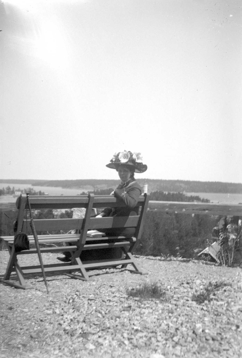 En kvinna med stor hatt med blommor sittande på en bänk.
