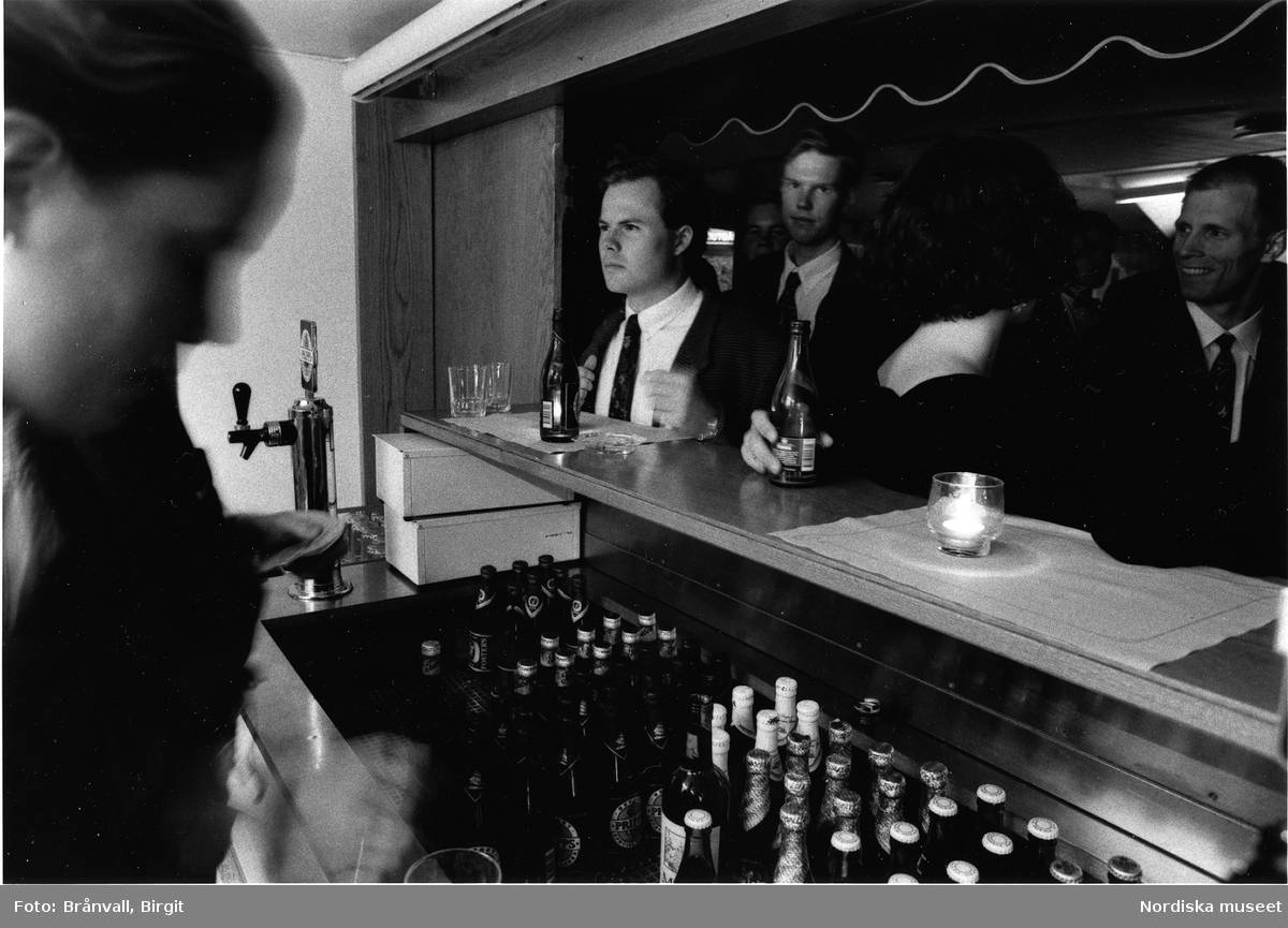 "Phösning", "nollning", för studenter till Flygsektionen vid Kungliga Tekniska högskolan i Stockholm 1993. Serveringspersonla vid bar, dukade bord för festmåltid. Scenframträdande.