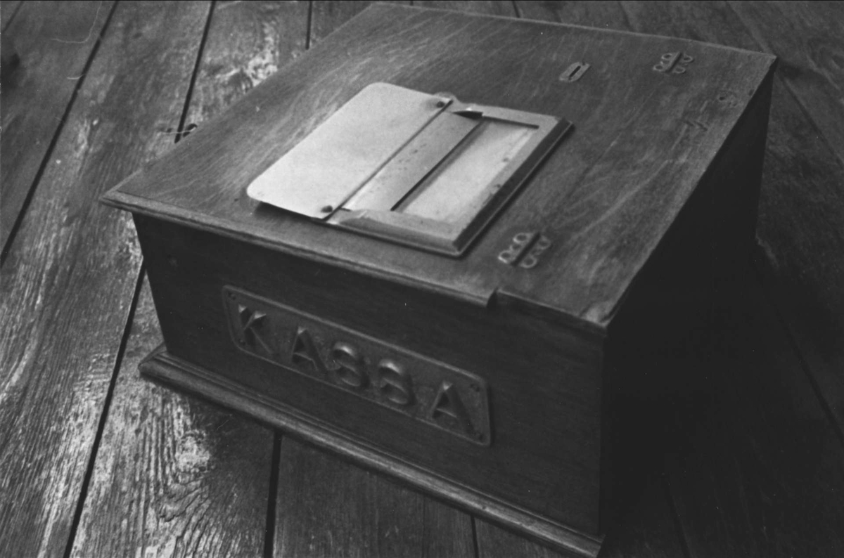 Kassapparat av trä med pulpetställt lock. Utdragbar låda för pengar, anordning för utskrivande av kvitton samt ringanordning. På kassaapparatens högra sida en gjutjärnsplatta med texten KASSA i relief.
