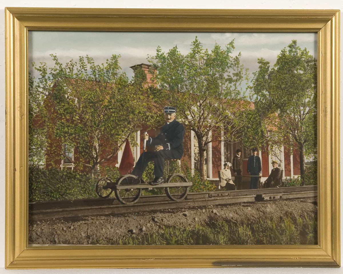 I förgrunden ett järnvägsspår, en stins sitter på en dressin. I bakgrunden en byggnad målad i rött med vita snickerier. Tre män och en kvinna befinner sig framför huset. Bilden färglagd i grönt, blått, rött och vitt.