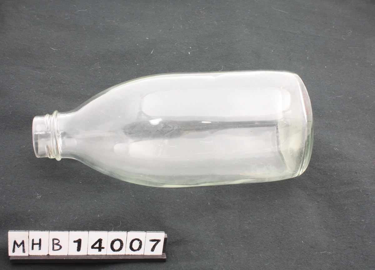 Melkeflaske i blankt glass. Bunnen er sirkulær, og flasken minker i diameter opp mot halsen. Øverst er gjenger til kork.