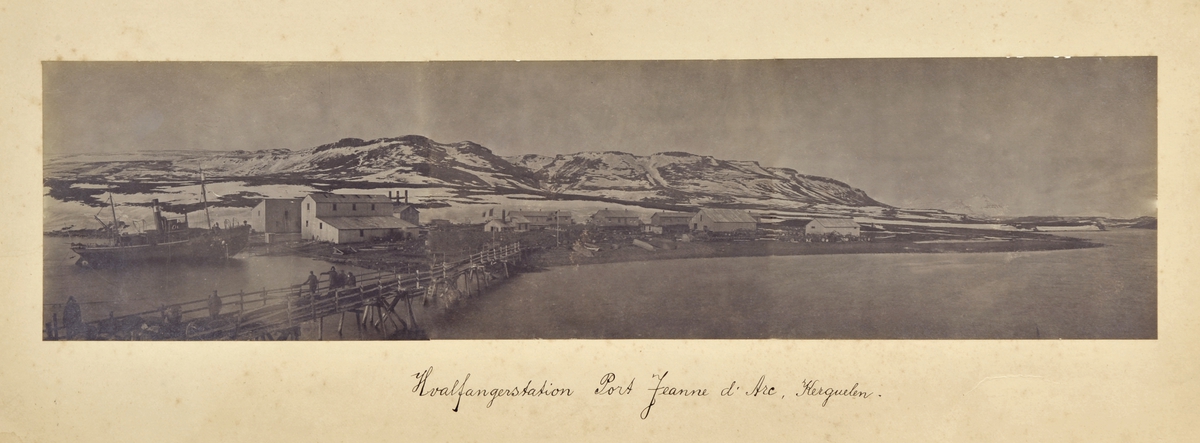 Panorama av 3 bilder. Til venstre hvalbåt på slipp, bygninger, lang brygge i forgrunnen, delvis snødekte fjell i bakgrunnen