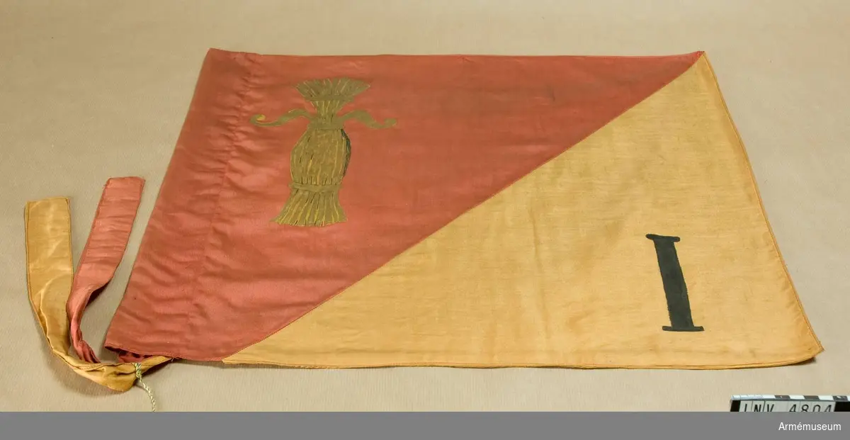 Mått: 540 x 440 mm.Wasa grenadjärbataljons s k riktflagga, Finland. Duken i  bomullssatin är tvärdelad med Övre delen röd och den undre  delen gul. Flaggan är sydd i dubbelt tyg, tryckt lika på båda  sidor. I det röda fältet visas Wasakärven och i det undre en  romersk I:a. Vid vänster sida en slå och band av tyget i rött  och gult för flaggans fästande vid stång.