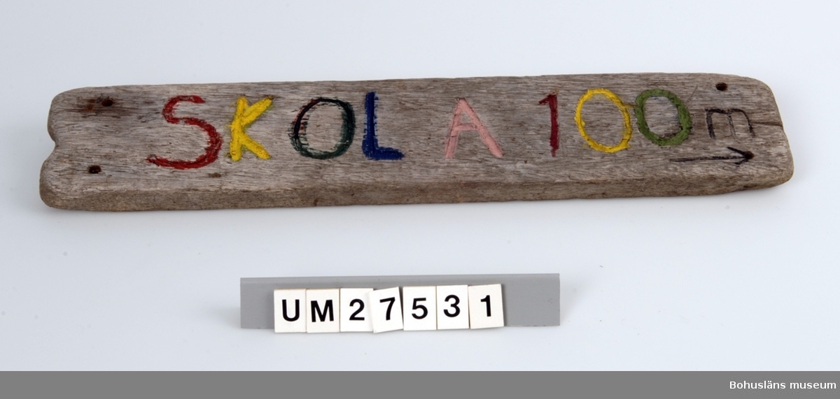 Vägskylt tillverkad av elev vid Åstols skola, Tjörns kommun.
Vägskylt tillverkad av en bit drivved. På framsidan inhuggna texten SKOLA 100 m, målat i blått, rött , gult och grönt samt en pil som pekar åt höger.  Borrhål för skruvar.

Föremålet visades i utställningen Moderna skärgårdsbor på Bohusläns museum 2002.
Utställningstext:
Vägskyltar
Nio skyltar som vägvisare på Åstol. Från affären till rökeriet är det 375 meter och från skolan till badplatsen är det 200 meter. Skyltarna berättar om en liten ö med många mötesplatser. Här finns affär, bibliotek och skola - restaurang, lekpark och flera församlingar. En tätbebyggd ö omgiven av havet, med skolan i centrum - så viktig för barnfamiljerna och åretruntboendet. Skyltarna är gjorda av elever i årkurs 4 - 6 på Åstols skola.

Skylten ingår i insamlingen i projektet Moderna skärgårdsbor, UM27511 - UM27595 och kommer från Åstol. 
För övrigt material från Åstol, se
UM27524-UM27538 och UM27566-UM27594;
UM27524 Tjörn Runt-teckning
UM27525- Vägskyltar, 9 st
UM27533
UM27534:1-2 Eternitplattor, 2 vita refflade 
UM27535 Eternitplatta, 1 vitmålad slät
UM27536:1-2 Eternitplatta, 2 gröna
UM27537 Cykelkärra
UM27538 Matjordsäck, 2 st (omärkt)
UM27566- 28 barnteckningar över Åstol, indiv. inv.nr.
UM27593
UM27594 Skylt frikyrkan med gudstjänstaffisch
Arkivet 9 uppsatser från skolbarnen i samband med teckningarna.

För information om projektet Moderna Skärgårdsbor, se UM27511.

Litt: Sjöholm, Carina. Moderna skärgårdsbor i gammal kultur. Skrifter utgivna av Bohusläns museum och Bohusläns hembygdsförbund nr 73. Bohusläns museums förlag. 
Avsnittet om Åstol s. 17 - 45..