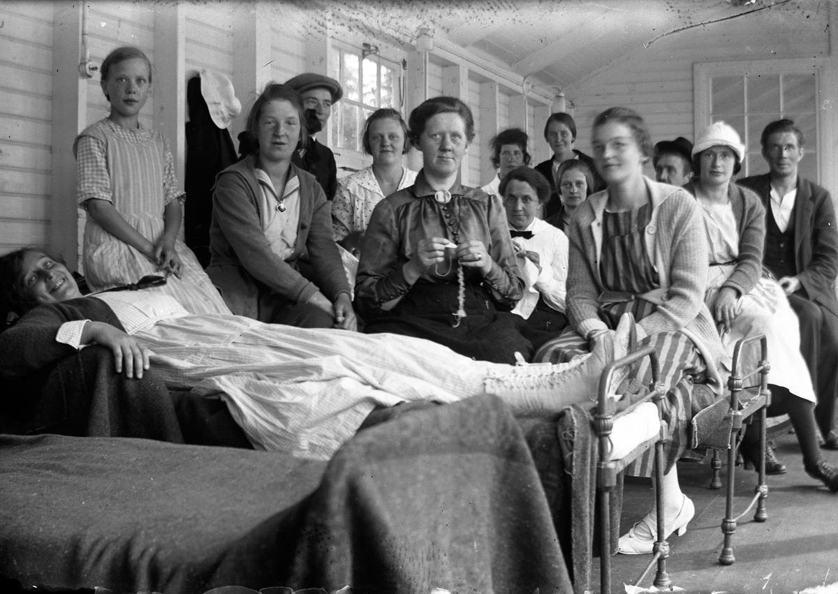 Tio kvinnor, tre män och en flicka varav en liggande och resten stående respektive sittande på eller vid en rad sängar på en träveranda på Lungkliniken i Eksjö.
Fotografens anmärkning: "Grupp i Hallen".