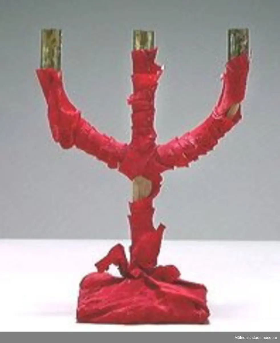 En s,k altarljusstake  för 3 st ljus, med mässinghylsor så som ljushållare, klädd med rött kräpp-papper. Ingår i en samling från Holtermanska daghemsverksamheten.