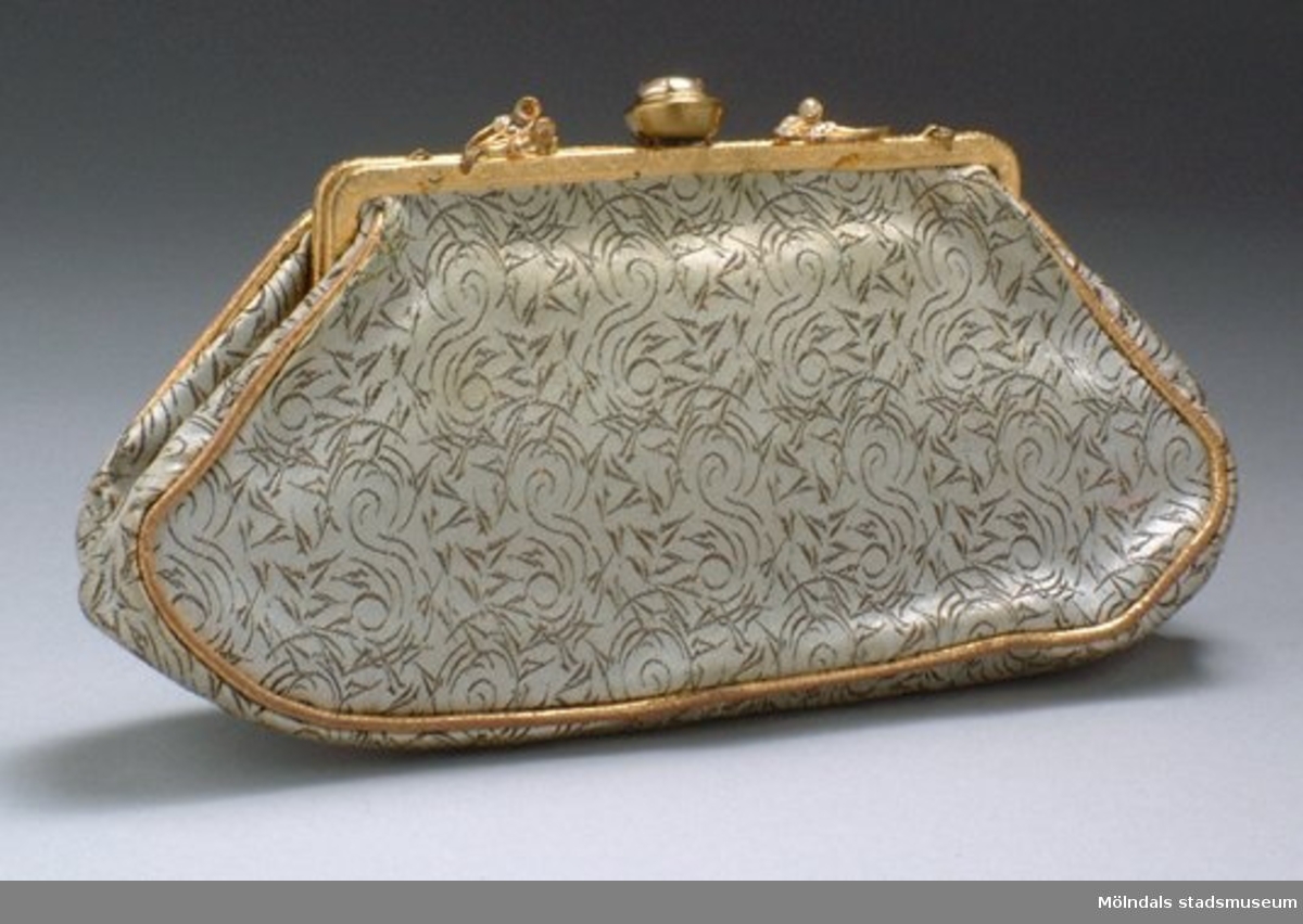 Liten oval handväska med "portmonnä" öppning. Den är vit och är guld/brun mönstrad. Öppningen är guldfärgad och har stenar som utsmyckning. Har troligtvis haft ett axelband, men är borta.Måtten:Bredd 220 mm, höjd 120 mm.