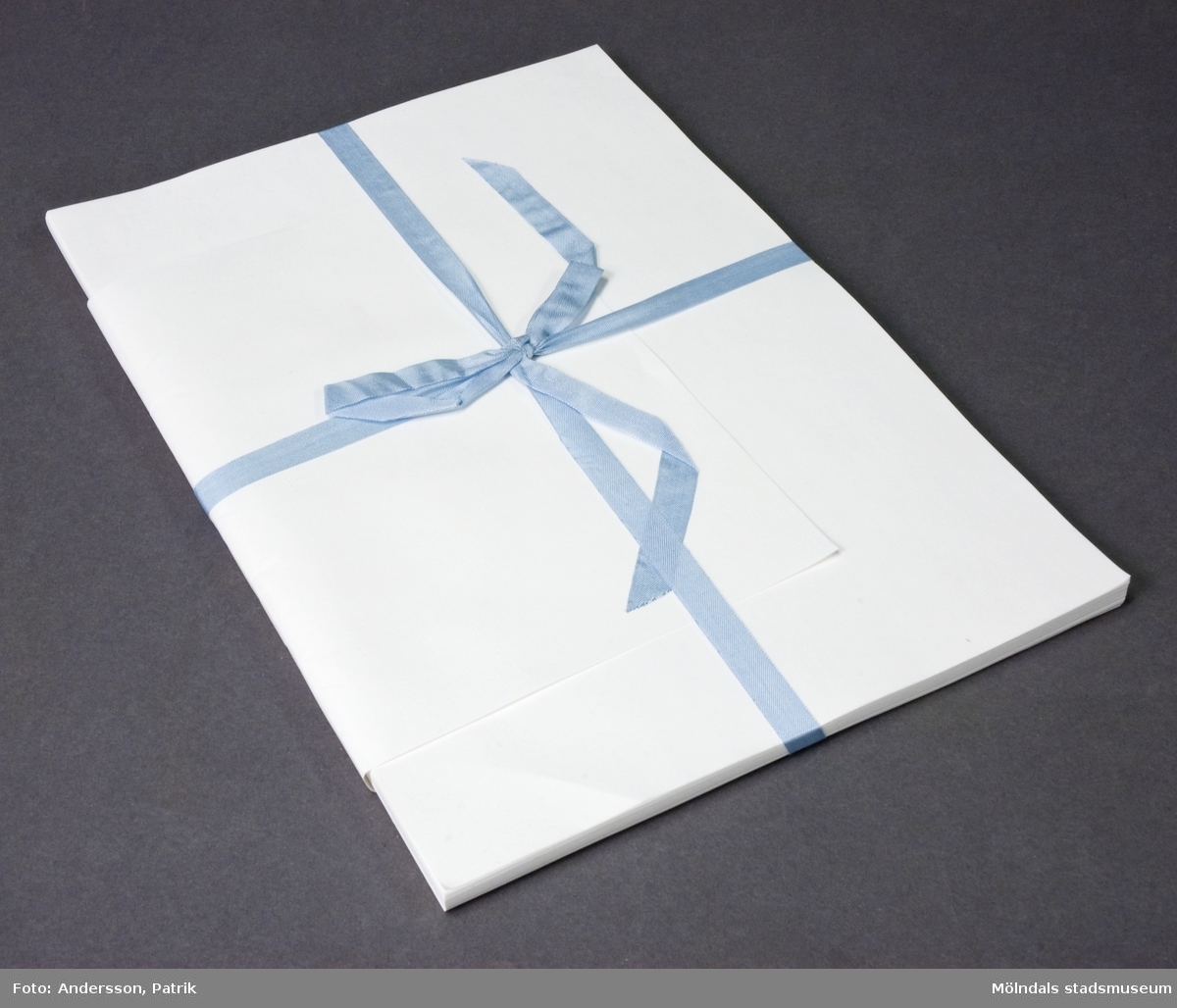 74 ark papper med linnestruktur, varav ett vikt runt de övriga, ombundna med ett blått sidenband. Bakerst i bunten finns ett vitt pappark med texten "MultiArt Silk 240 g/m2".Pappret är tillverkat på Papyrus, Mölndal. 
