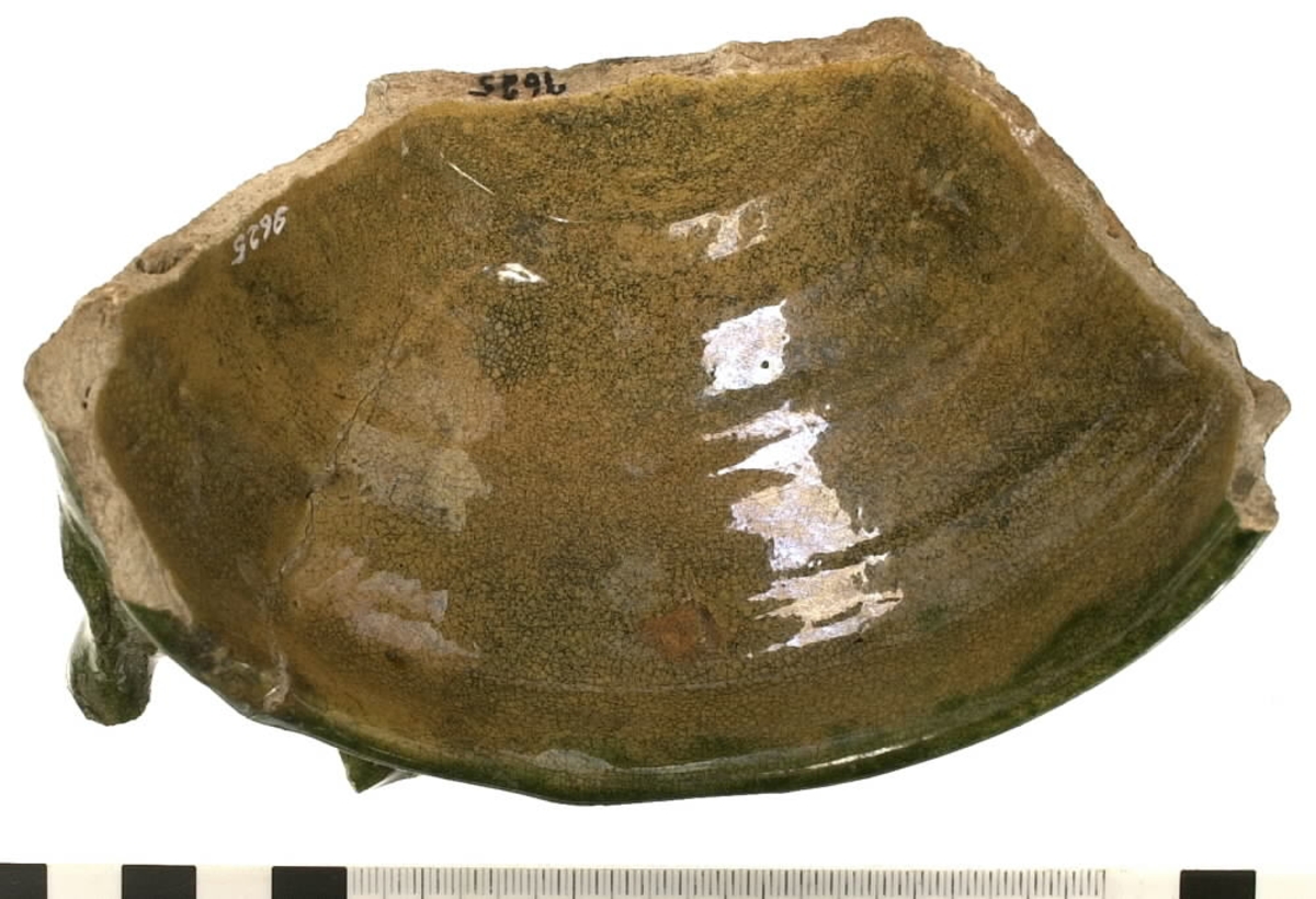 Del av en keramikskål. Skålen är tillverkad i vitbränt lergods. Skåldelen är försedd med en avbruten hänkel och utgör ena halvan av den ursprungliga skålen. Utsidan är glaserad med en grön glasyr och insidan med en senapsgul glasyr.