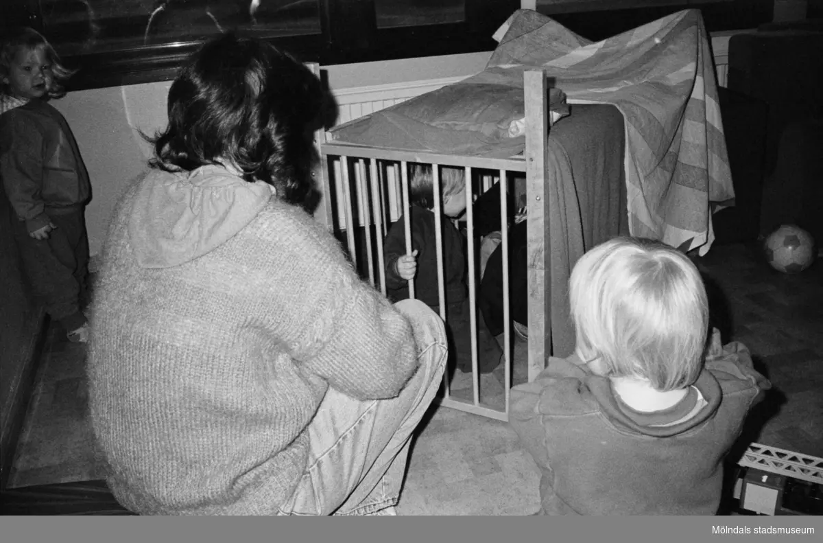 En förskollärare vid Katrinebergs daghem, hjälper barnen att bygga koja inomhus med hjälp av filtar liggandes över en spjälsäng. Två barn sitter utanför och två barn sitter i "kojan". I bakgrunden ser man en fotboll.