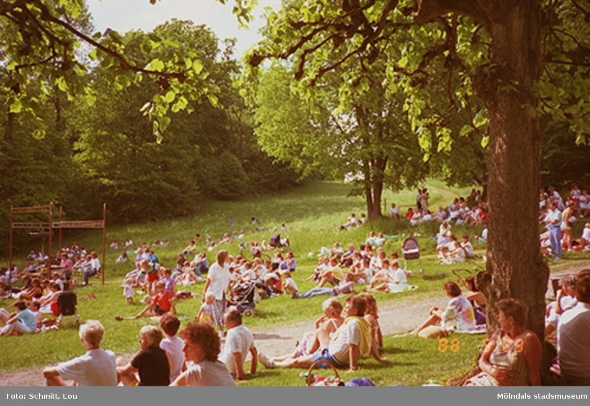 Människor som sitter i gräset och lyssnar på Lasse Berghagen (ej i bild) som sjunger på en tillfälligt uppbygd scen. Gunnebo slotts park, 29 maj 1988.