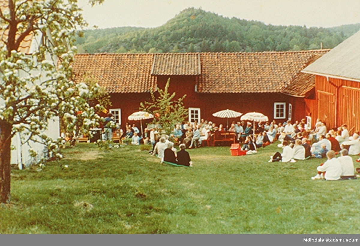 Människor sitter i gräset och på bänkar utanför Börjesgårdeen/Lindome Hembygdsgård. De lyssnar på ett uppträdande. Okänt årtal.
