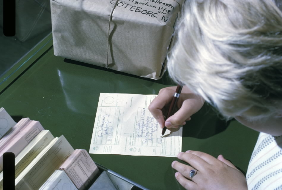 Seriebild 28 M. Lisa Brännfors fyller i "Innehåll" på adresskort.
Paketet ligger färdigt bredvid. Hon står vid en skrivpulpet. Intill
finns ett ställ med blanketter.