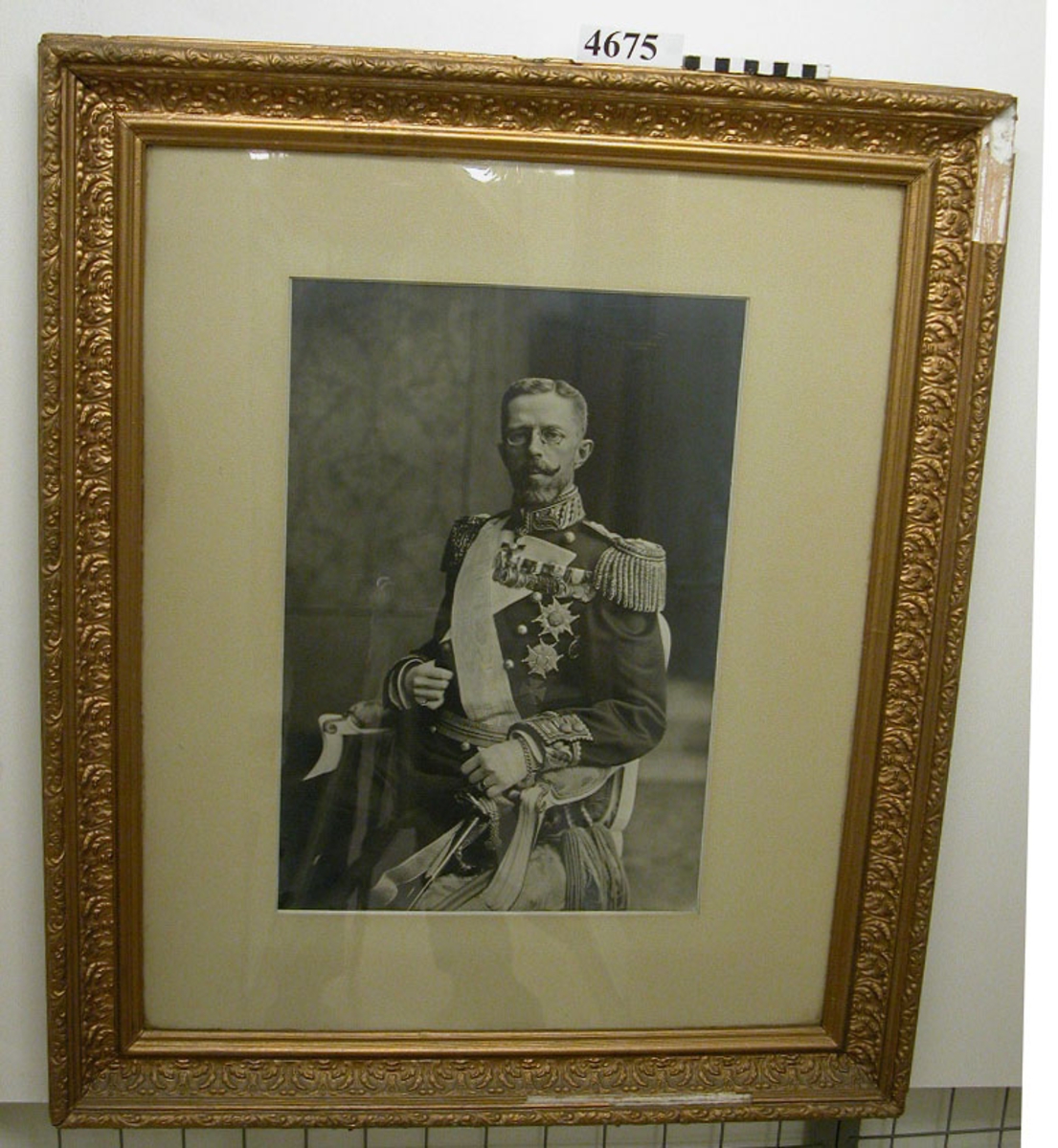 Fotografi av H.M. Konung Gustav V. Ramen av trä och gips med kunglig krona på överkanten, bronserat i gult. Fotografiet märkt på baksidan med "Örnen" samt kronstämplar.