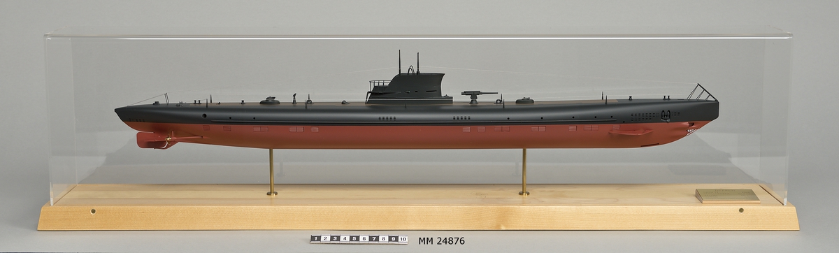 Ubåtsmodell Draken i monter. Modell av alträ med detaljer av mässing, målad med cellulosafärg. Rött och svart skrov. Monter av plexiglas på träplatta. Mässingsbricka i montern med uppgifter om modellen.