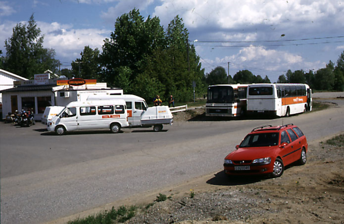 Lufthavn, 2 busser og 3 andre kjøretøyer parkert på parkeringsplass. 2 bygninger i bakgrunnen.