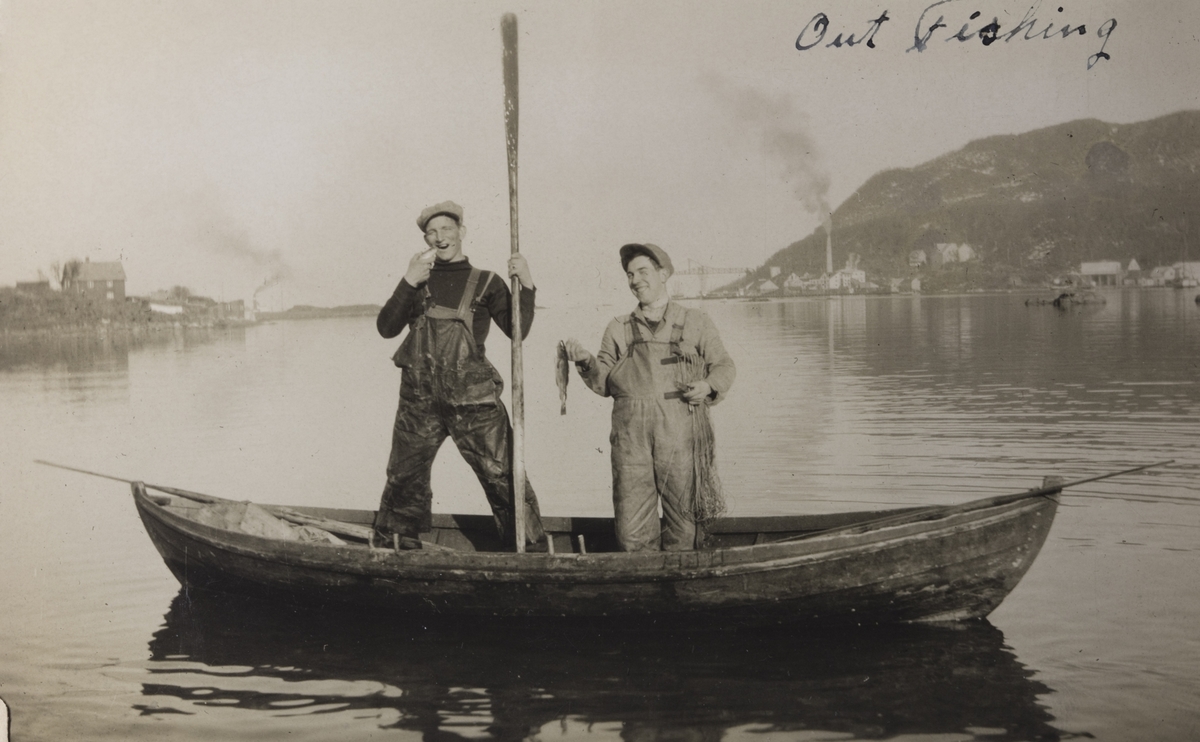 To menn i liten robåt poserer stående i båten med hver sin fisk. I bakgrunnen Mercur sildoljefabrikk. Mannen til venstre er Haakon Iversen.