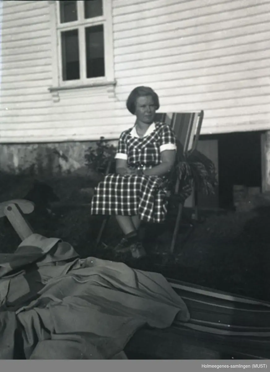 En ung kvinne i sommerkjole og hatt sitter i en solstol utenfor et bolighus. Antatt samme sted og tidspunkt på ST.K.HE 2010-011-0091 til -0094.