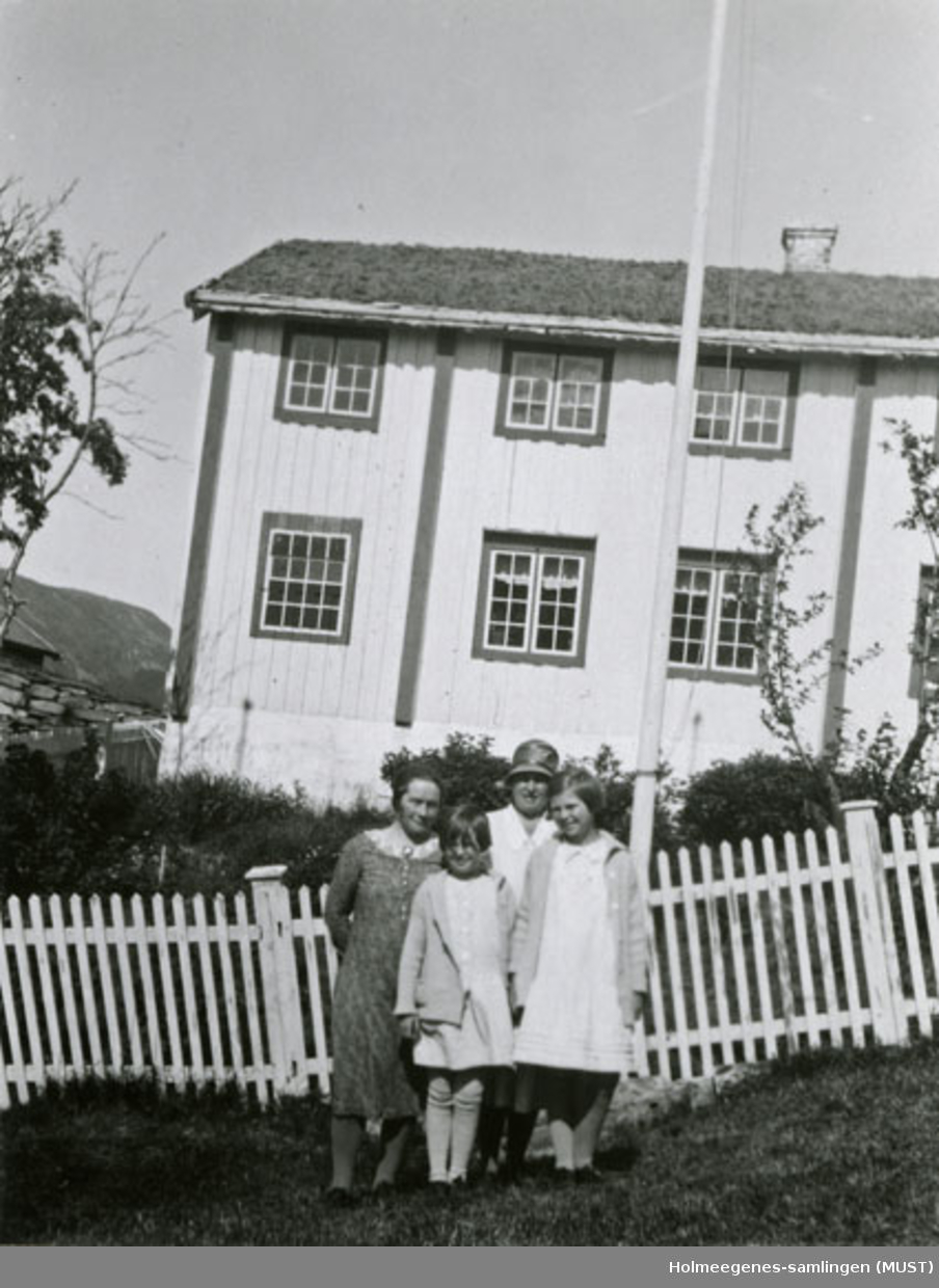 To kvinner og to jenter i sommerkjoler står foran et hvitmalt tregjerde, i bakgrunnen et bolighus med to etasjer. Se også ST.K.HE 2007-011-0111 til -0126.