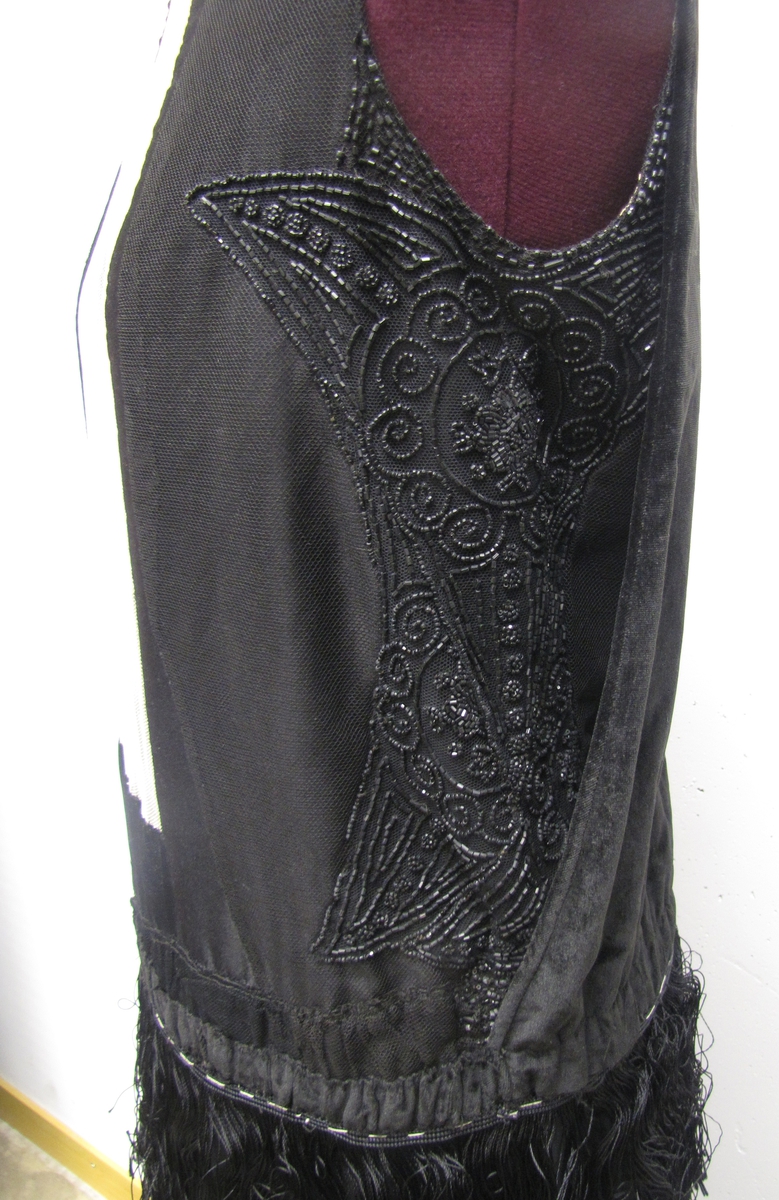 En svart ärmlös klänning med frans och pärlbroderier.

Kjolen har 5 rader med silkefransar.
 Pärlbroderier på svart tyll på  livets sidor fram. Mitt fram finns vit tyll med spets infälld.
 Livets baksida består av sammet.