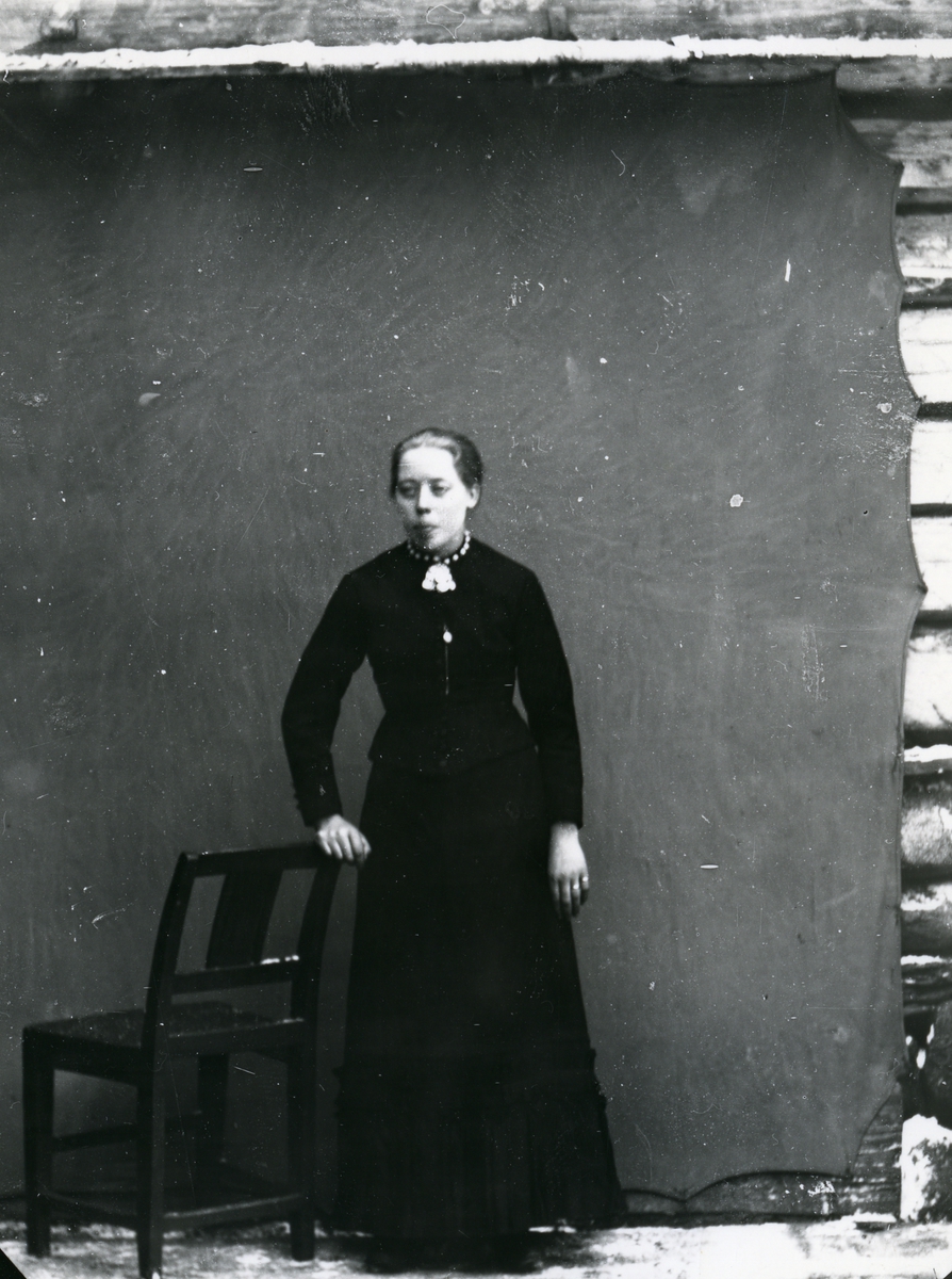 Kvinne i helfigur, stående ved stol foran lerret