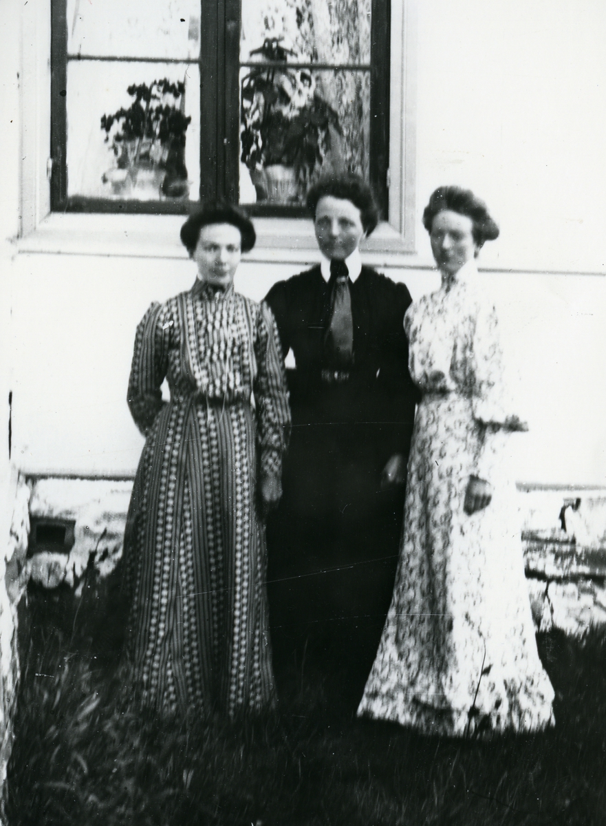 Tre kvinner i helfigur, fotografert i hushjørne foran vindu med planter innenfor