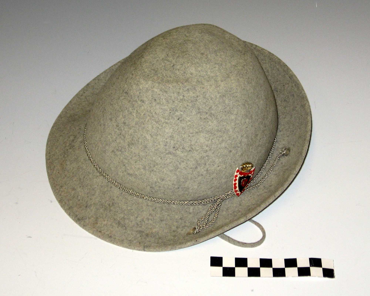 Hatt.Liten lysegrå filthatt, østeriksk/bayersk type, med tvinnet snor rundt pullen (innerst på bremmen) og merke/emblem (etterligning av noe). Hatten ble brukt til pent (17. mai) - av en gutt på 4 år.