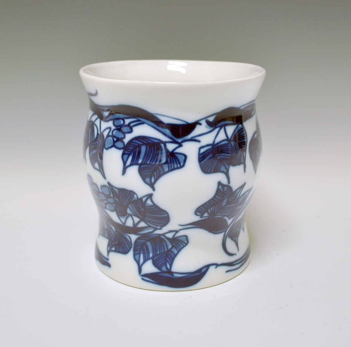 Vase i porselen. Bølget form, sirkelformet fot og oval åpning på toppen. Hvit glasur. Blå underglasurdekor: Grener med blader og drueklaser.
Kunstner: Unni M. Johnsen.