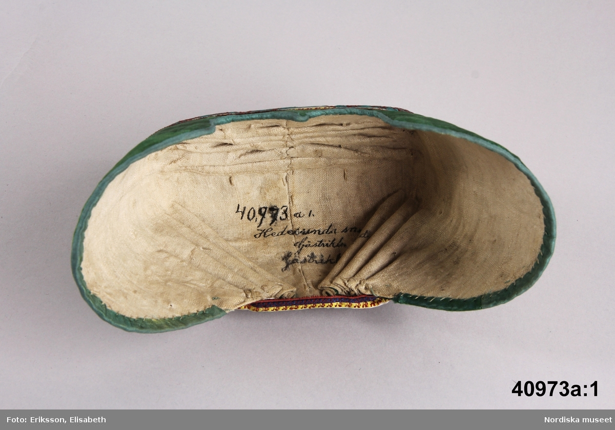 Huvudliggaren: Dräkten "begagnades till början av 1840-talet, linnet av senare årtal."
a.1-2 bindmössa m. stycke
b. tröja längd 35 cm, ärml. 46 cm
c. överdel
d. förkläde
e. kjol
f. 1-2, skor
g. 1-2, bindmössa m. stycke

a.1 BINDMÖSSA av grön sidenatlas med broderi i tambursöm med silke i gult, vitt, rosa, rost, svart och ljusgrönt. Kantad med blågrönt sidenband. Mittsöm över hjässan samt materialskarvar framtill vid sidorna. I nacken 4 och 4 mot mitten lagda veck. Foder av ljus linnelärft. Mönstrat sidenband runt kullen i rödbrunt, lila, gult och vitt.
/Berit Eldvik 2005-09-08