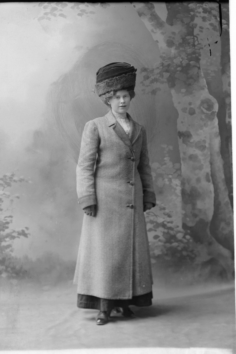 Studioportrett av en kvinne i kåpe og med hatt på hodet.
