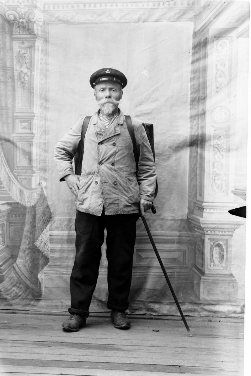 Studioportrett av en postmann med ryggsekk, uniformslue og vandrestav.