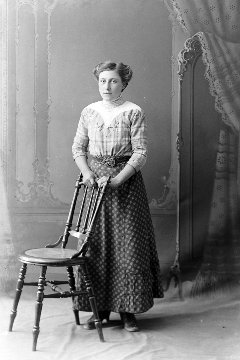 Studioportrett av en kvinne med en stol foran seg.