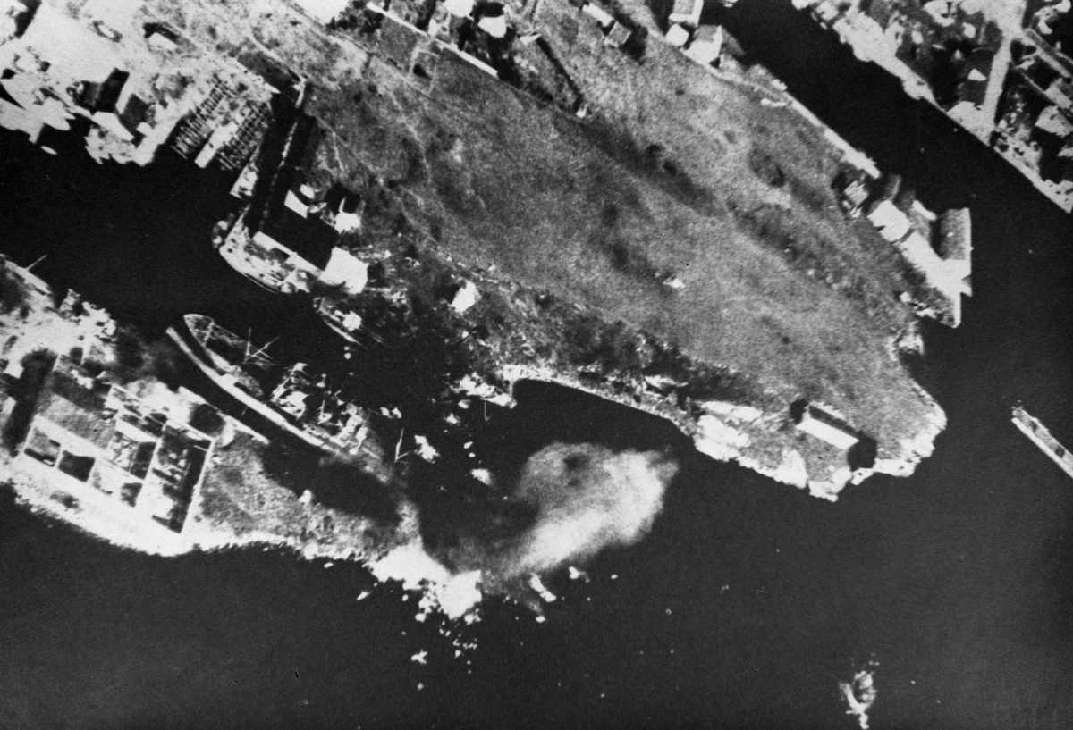 Luftfoto av skip i brann. Skipet ligger i sundet mellom to øyer.
Sydspissen av Risøy med "slippen" og HMV.