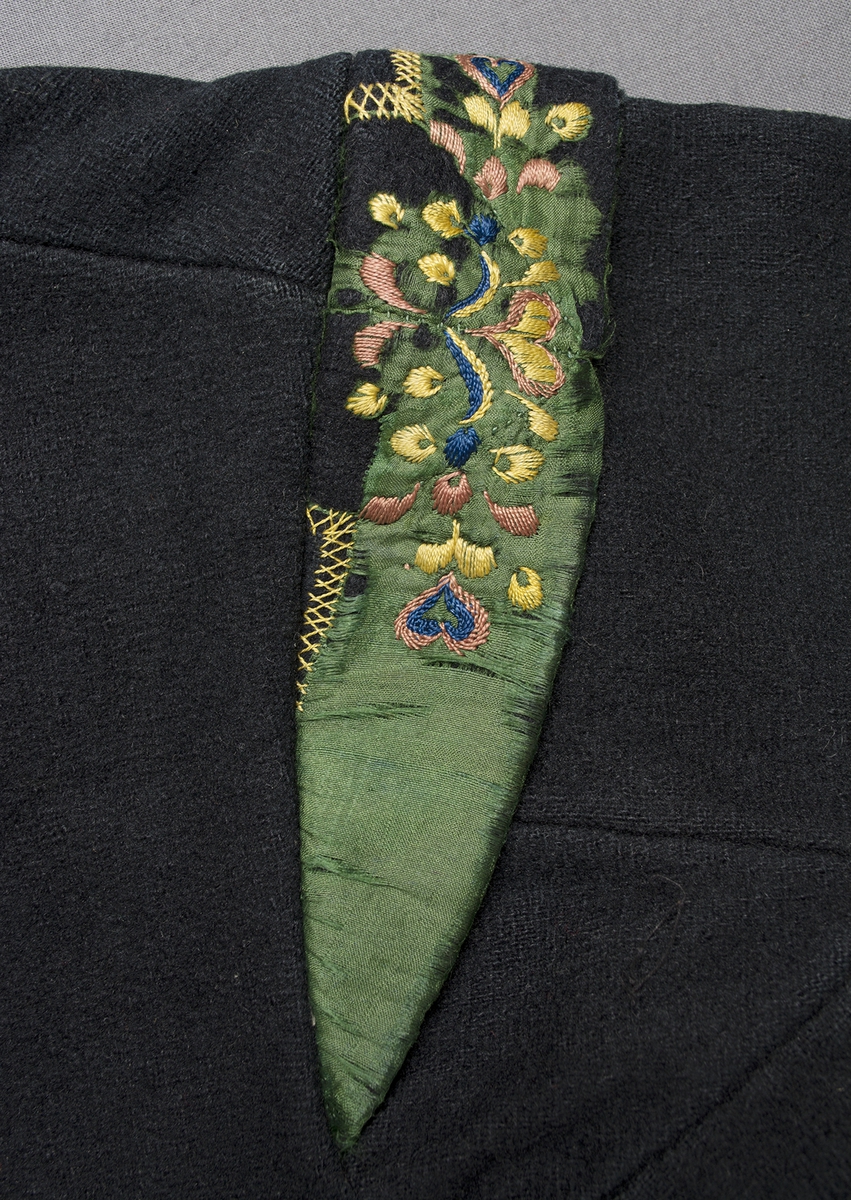 Mörkblå vadmalströja garnerad med gröna sidenband och broderier. Tröjan har rund ringning fram, skört bak och axelkarm på ärmen. Tröjan är dekorerad med 50 mm breda gröna sidenband längs halsringning och ringning fram, längs nederkanten på framstycke och ärm samt på ärmkarmen. Ovanför ärmsprundet har ett kryss av banden sytts på, ovanför sitter en rosa "ros" i siden. Dekorativa broderier i plattsöm, kråkspark och flätsöm på och invid de gröna banden på framstycken, ärmar och ärmkarmar samt vid skörtflikarna bak. De fantasifulla broderierna i form av bland annat hjärtan och blommor är sydda med silke i gult, rosa, blått, gulaktigt vitt och grönt. Framtill knäpps tröjan på två ställen med en hyska och hake. Söm mitt bak med skörtflik nedtill, ytterligare två skörtflikar bak, en intill vardera sidan. Axelbredd: 100 mm. Axelkarmen är som bredast 40 mm bred. Ärmlängd: 480 mm. Ärmen är sydd i ett stycke, med en kil i ena ärmen. Ärmsprundet är 90 mm långt och knäpps med en hyska och hake. Tröjan är helfodrad med halvblekt handvävt tuskaftat linne, grövre i ärmarna.Märkt invändigt på ena framstycket med en påsydd tyglapp med texten: "N° 161 c.".