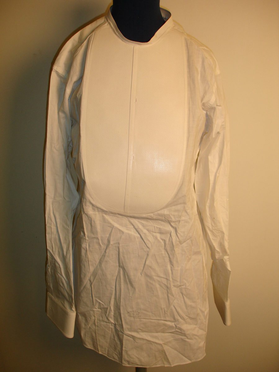 Forkle, med påsydd stivet bryststykke og mansjetter. Skjorten kneppes bak, er en trykk-knapp til lukking. Kan være skjorte til kjole og hvitt, eller skjorte til sykepleieruniform.