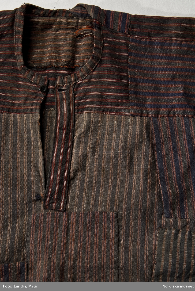 Skjorta, bussarong, arbetsklädsel, Uppland, Gräsö, 1900-talets början. Halvylle. 
Arbetsskjorta s.k. bussarong av hemvävt halvylletyg. Mörkblå bomullsvarp och inslag av ullgarn i mörkgrått och mörkblått med röda ränder och i vissa partier även en vit tråd. Traditionell skjortmodell med vidsydda ärmar med ärmspjäll (fyrkantig isättning) och ärmlinning. Skjortan är lappad upprepade gånger med totalt 19 lappar av olika randade tyger. Skjortan är maskinsydd, lapparna påsydda för hand, ofta på varandra. Den har använts ända fram till detta stadium av förslitning och blivit omsorgsfullt vårdad och lagad. Använd av fiskaren Karl Eriksson, på Gräsö, längst upp i Stockholms skärgård.