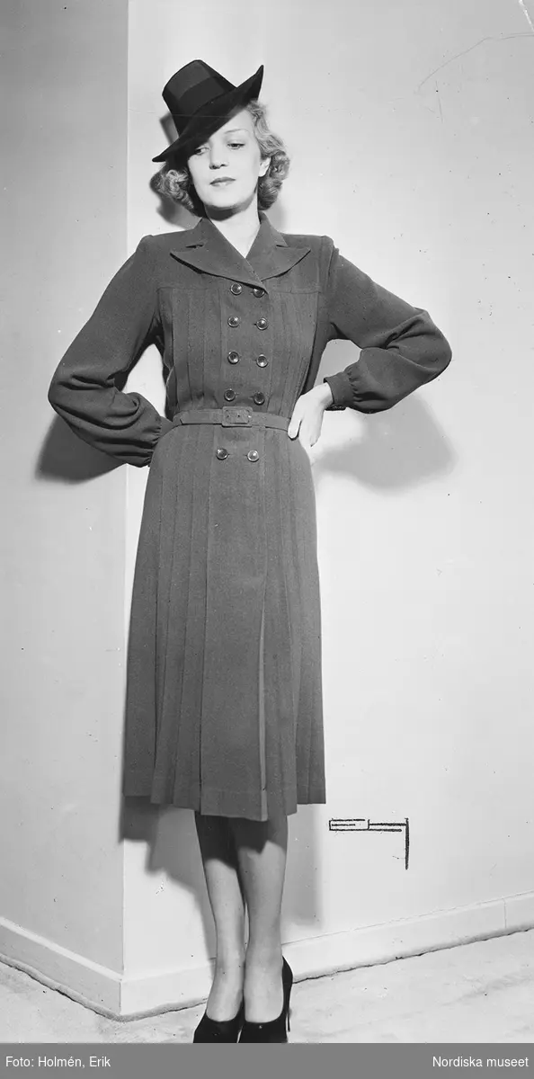 Modell i dubbelknäppt klänning, hatt och pumps.
