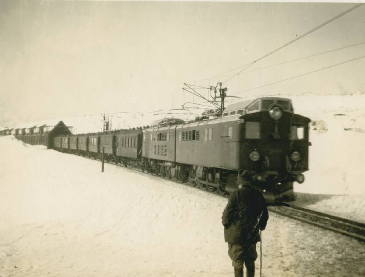 Lokomotivet er et elektrisk lokomotiv av type El.3  med  passasjervogner. utenfor snøoverbygg. 
Mann med hatt og stokk.