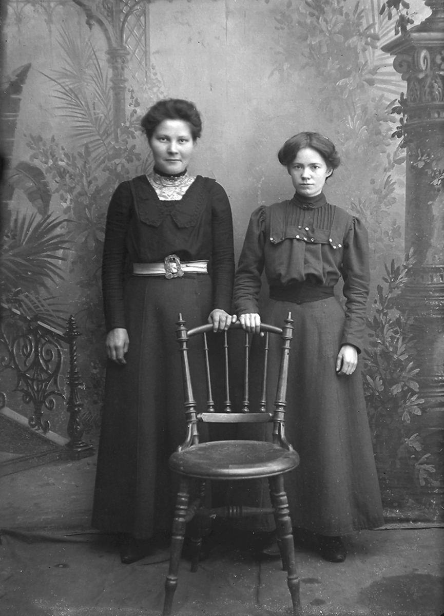 Portrett. To damer, begge står bak en pinnestol og begge har sin ene hånd på skulderbrettet på stolen. Begge har på seg kjole med belte. Bildet er tatt på Reine i Lofoten.