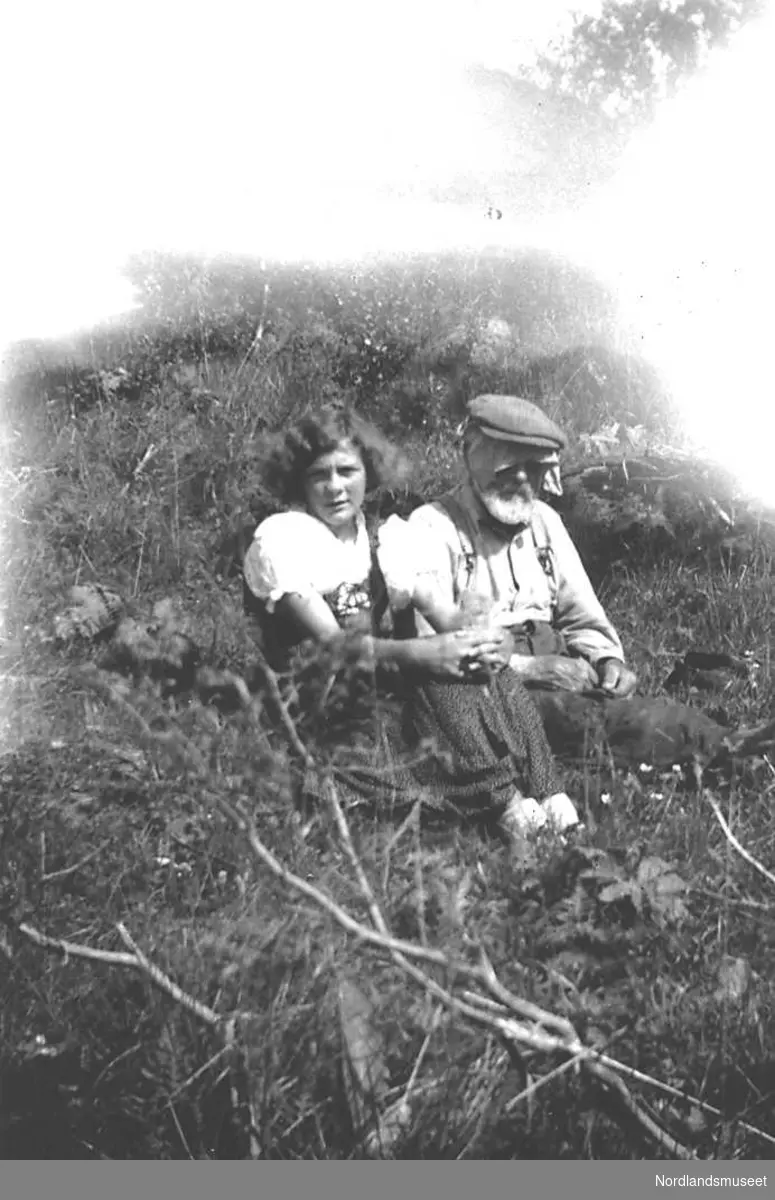 Portrett av ei ung jente , Ragna Larsen, og en gammel mann, Almeier Mikkelsen,  som sitter på bakken en  sommerdag ca. 1934. Det må være varmt, for Almeier har knyttet et stort lommetørkle rundt hodet under sixpencelua. Laksådalen, Gildeskål.