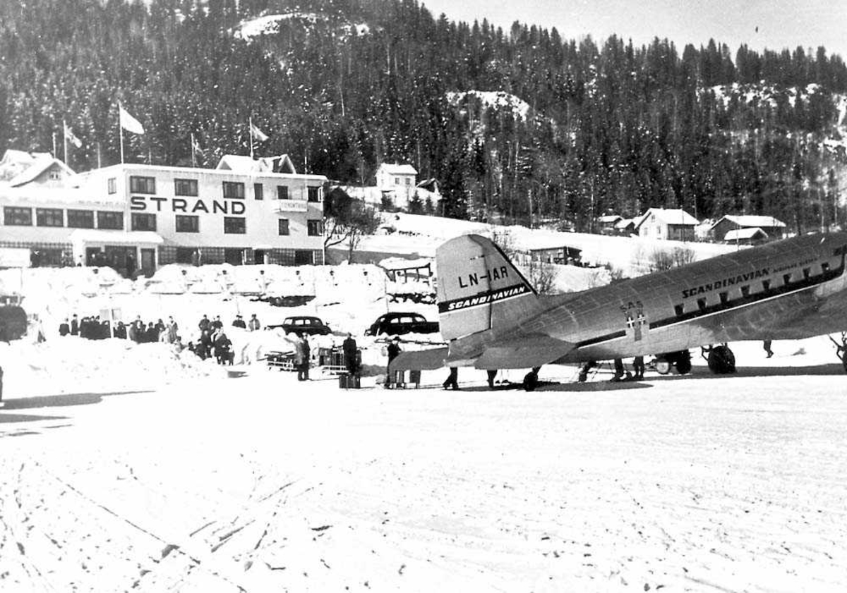 Ett fly på bakken, Douglas DC-3 / C-47 Dakota LN-IAR fra SAS. Flere personer ved flyet og i bakgrunnen. Bygninger og fjell i bakgrunnen. Snø på bakken.