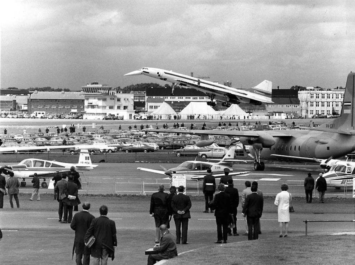Flystevne. Ett fly i luften like over rullebanen, Concorde 01. Flere andre fly på bakken. Flere personer. Bygninger i bakgrunnen.