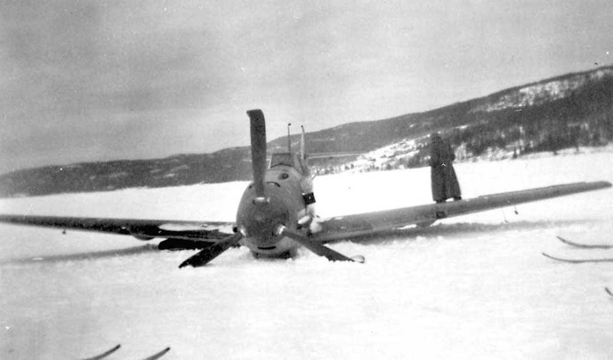 Ett havaret fly som ligger ute på et snødekt vann, Messerschmitt Me 109. En person står bak flyet. Fjell i bakgrunnen.